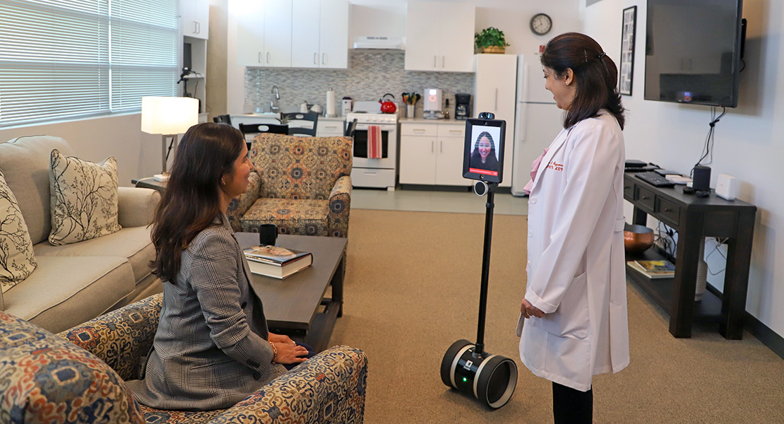Dr. Carina Katigbak and Dr. Seema Aggarwal in a clinical training room stand beside a telepresence robot showing a colleague's face on a tablet screen; one wears a white lab coat and the other a gray blazer.
